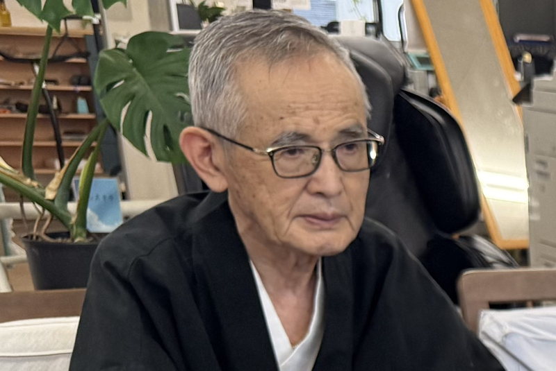 藤井薫。和装で椅子に座り、机をはさんで話を聞いている。